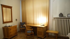 A room with a wooden desk, two chairs, a small bench, a radiator, and a mirror on the wall.