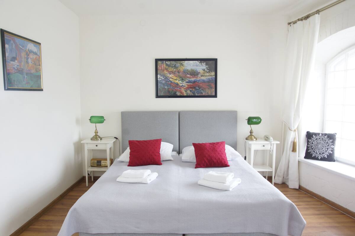 Een netjes opgemaakt bed met witte lakens en twee rode kussens, een nachtkastje met een lamp, en een schilderij aan de muur.