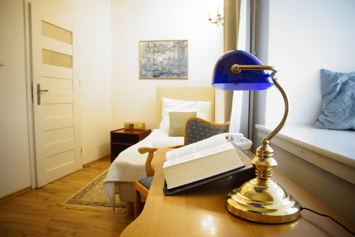 Een kamer met een houten bureau, een blauwe bureaulamp, een open boek, een bed met kussens, een nachtkastje, een schilderij aan de muur en een raam met uitzicht.