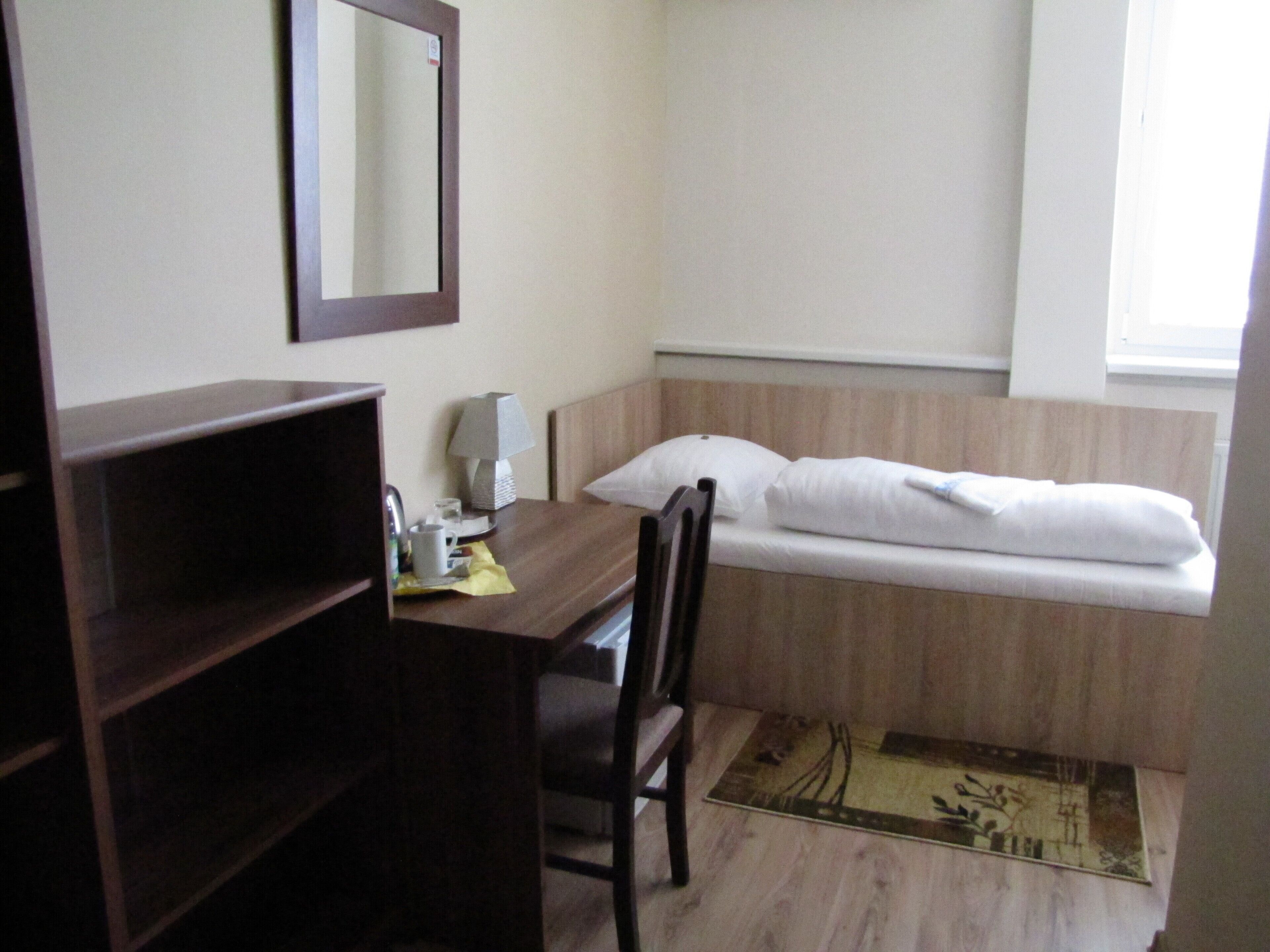 A hotel room with a wooden desk, a chair, a bed with white bedding, a bedside table with a lamp, and a window.