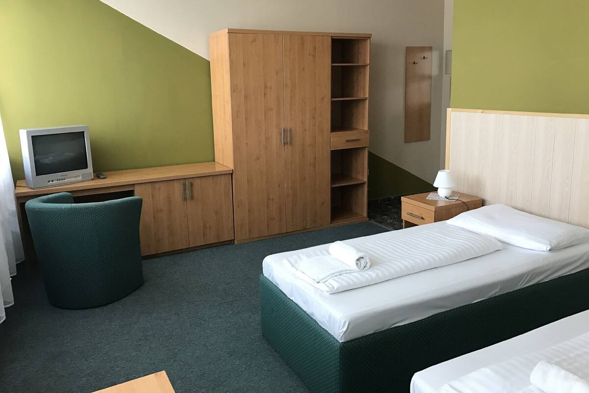 A hotel room with a bed, a desk with a television, a wardrobe, a chair, and a bedside table with a lamp.