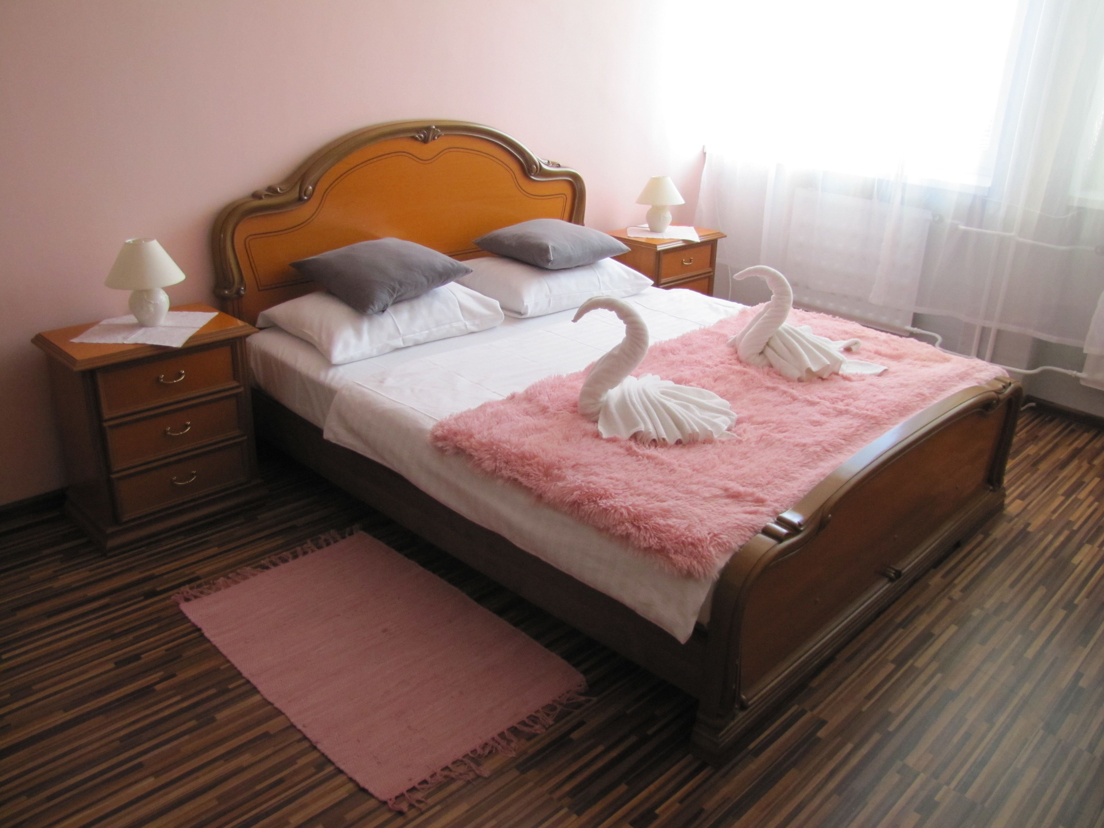 Una cama cuidadosamente tendida con un edredón rosa esponjoso y dos almohadas con forma de cisne. Hay una mesita de noche de madera con una lámpara y un cajón pequeño. La habitación tiene suelo de madera y una alfombra rosa.