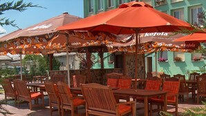 Un área de estar al aire libre con mesas y sillas de madera, cubierta por grandes sombrillas naranjas con la marca 'Kofola'.