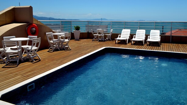 Zona junto a la piscina con sillones de descanso y mesas, un flotador salvavidas y vistas al mar.