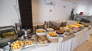 Disposición de buffet con diferentes opciones de comida, incluyendo pan, pastelería y postres, sobre una mesa rodeada de sillas.