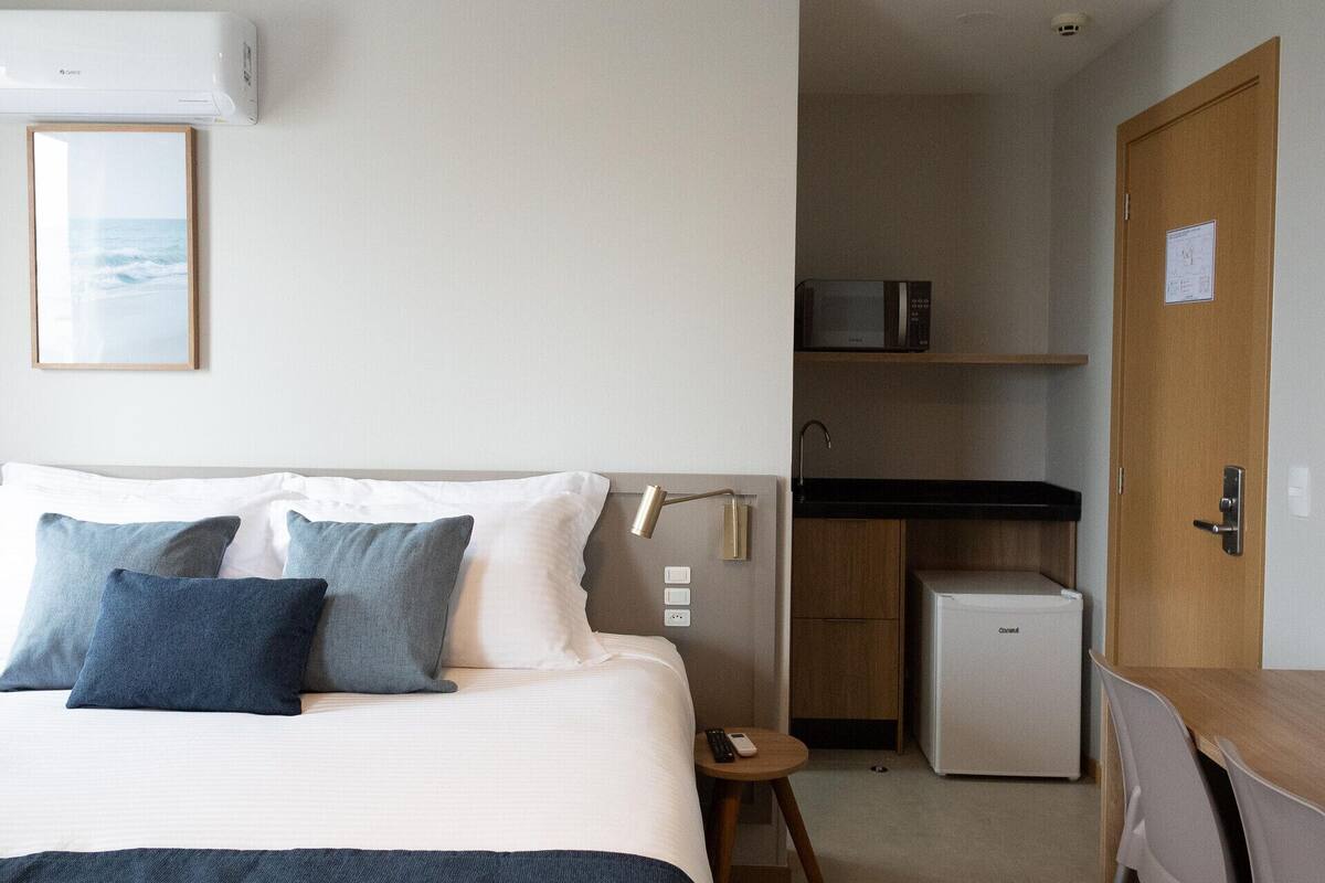 A hotel room with a bed, bedside table, desk, chair, microwave, refrigerator, and a small kitchen area.