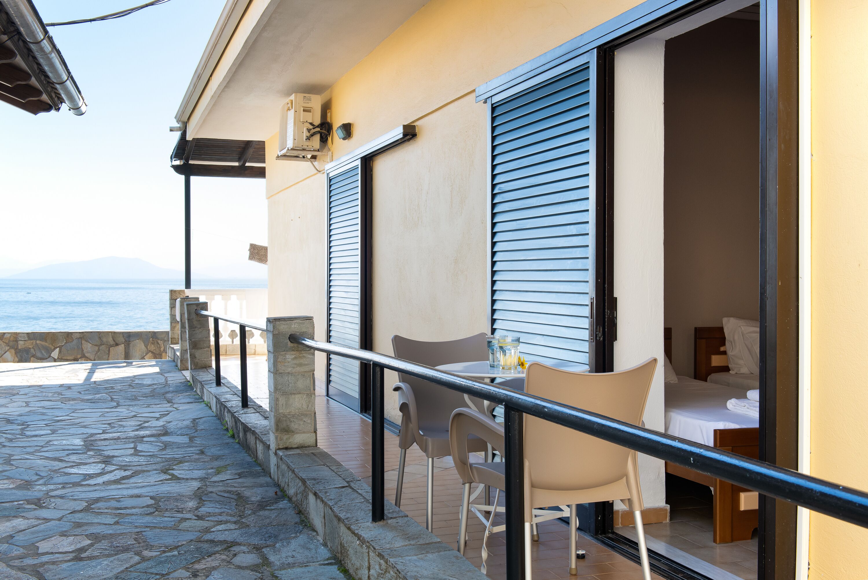 A balcony with stone flooring, a table with chairs, and a bed with blue shutters.