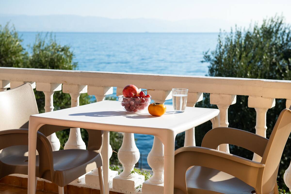 A table with a bowl of fruit and a glass on a balcony overlooking the sea.