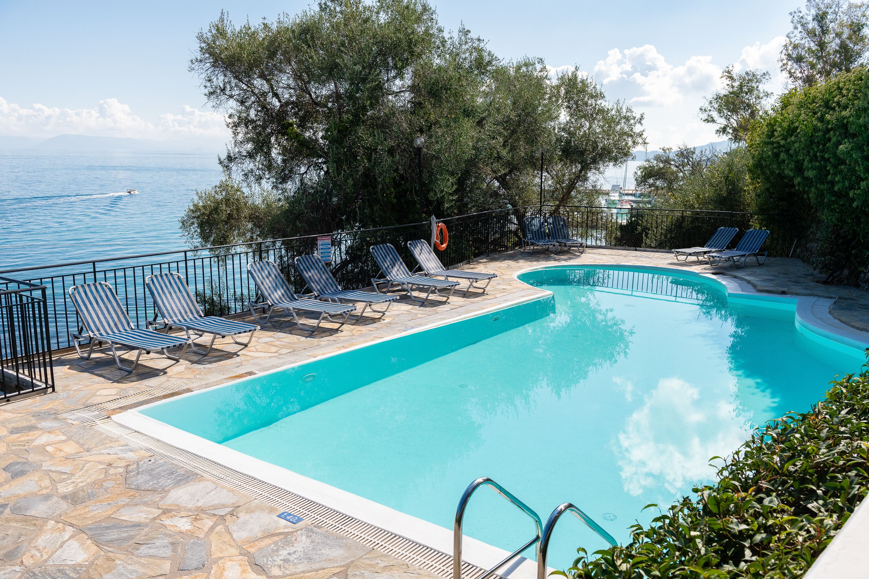A swimming pool with lounge chairs and a lifebuoy, surrounded by trees and overlooking the sea.