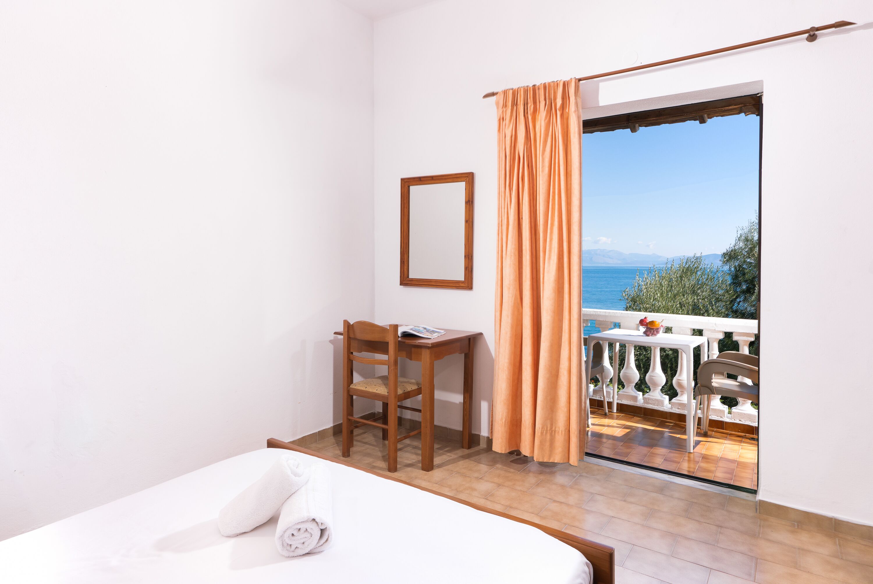 A hotel room with a bed, a wooden chair, a small table, and a view of the sea through an open balcony door.