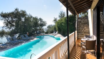 A swimming pool with lounge chairs and a table on a balcony with a view of trees and a building.