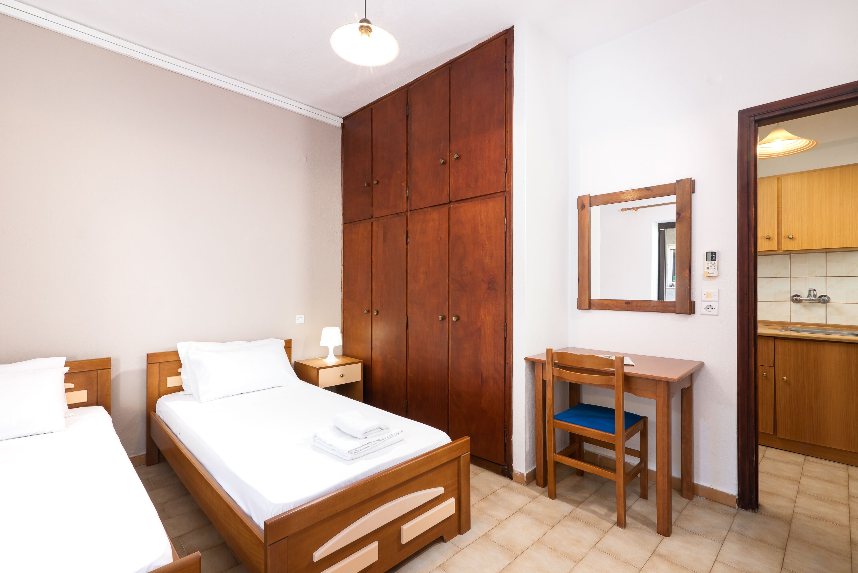 A bedroom with two beds, a wooden wardrobe, a desk with a chair, and a mirror on the wall.