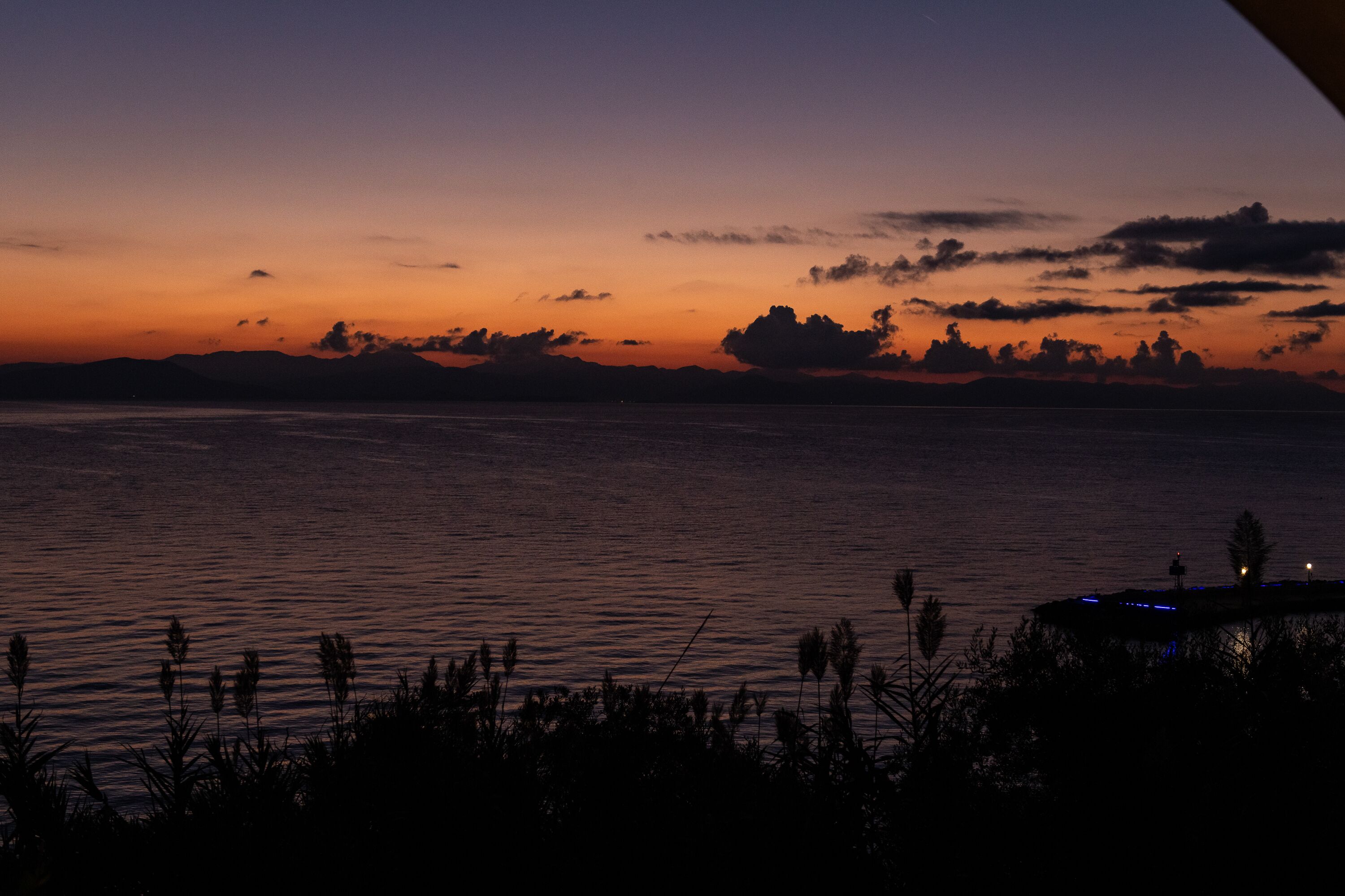 A sunset over a body of water with silhouettes of vegetation and a distant shoreline.