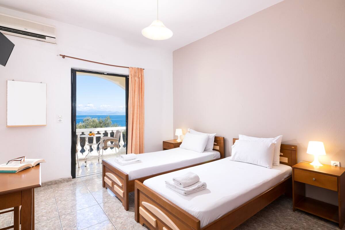 A hotel room with two beds, a desk, a chair, a balcony with a view, and a ceiling fan.