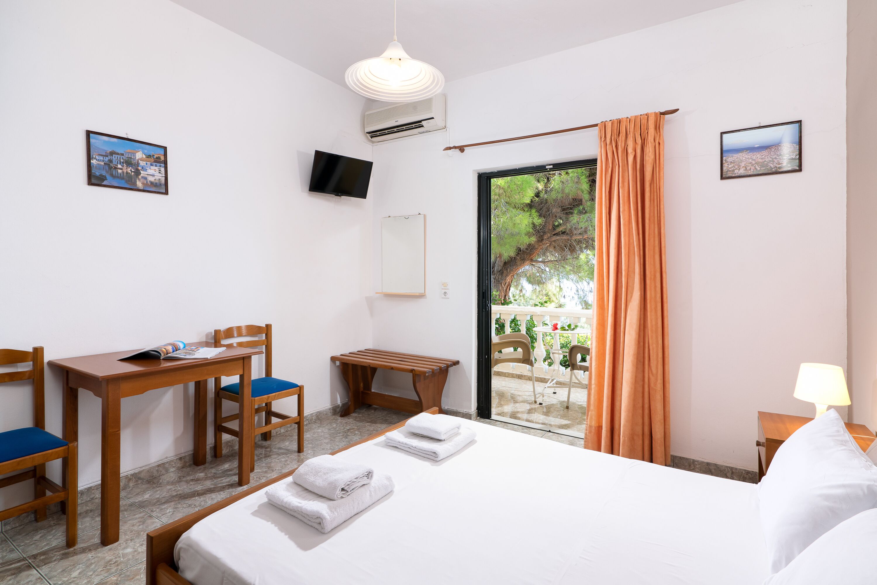 Quarto de hotel com cama, escrivaninha com cadeiras, televisão e varanda com vista para árvores e paisagem urbana.