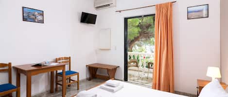 A hotel room with a bed, a desk with chairs, a TV, and a balcony with a view of trees and a cityscape.