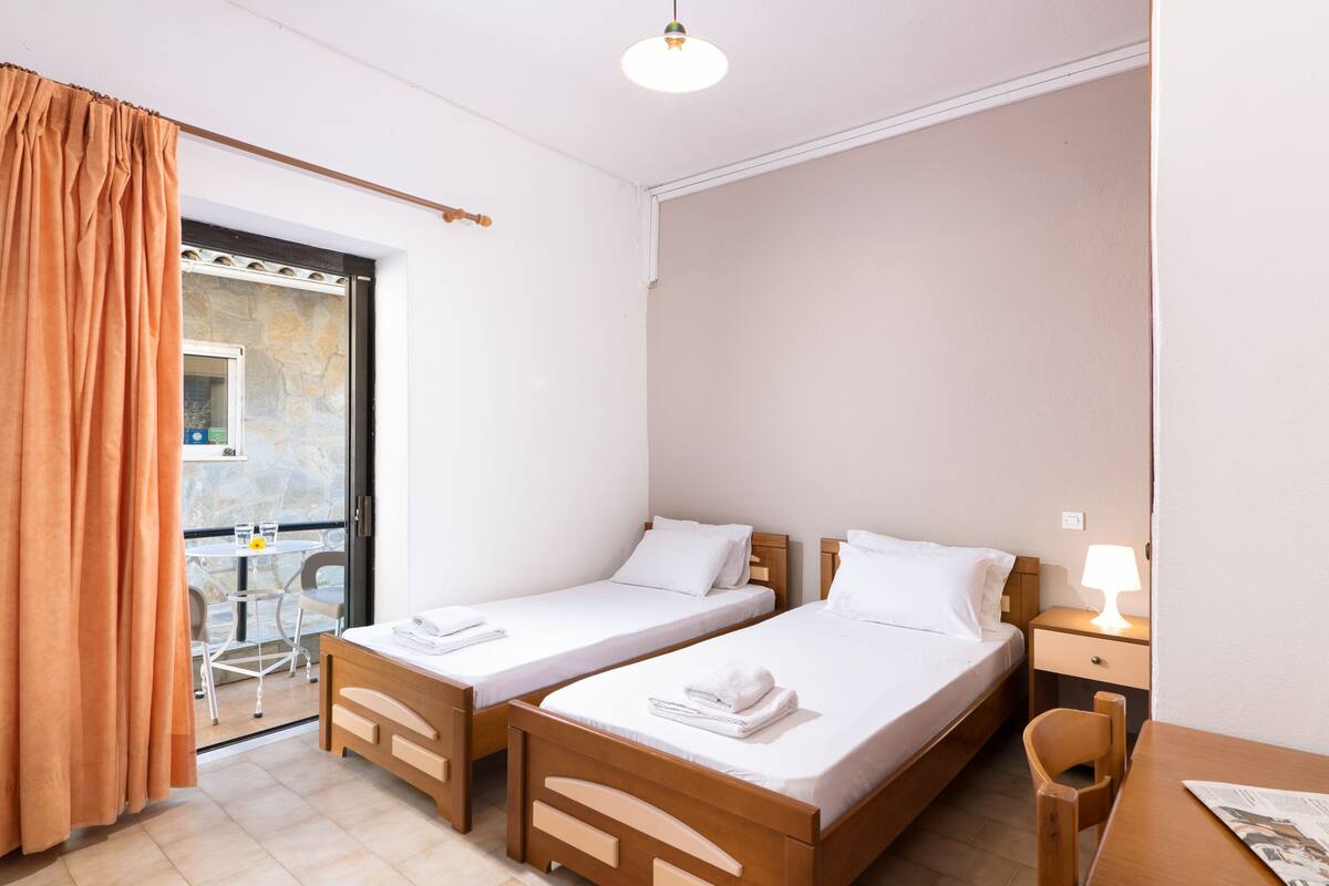 A room with two wooden beds, a small table, a chair, and a balcony with a table and chairs.