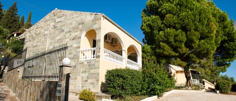 A stone building with a balcony, surrounded by greenery and a clear sky.