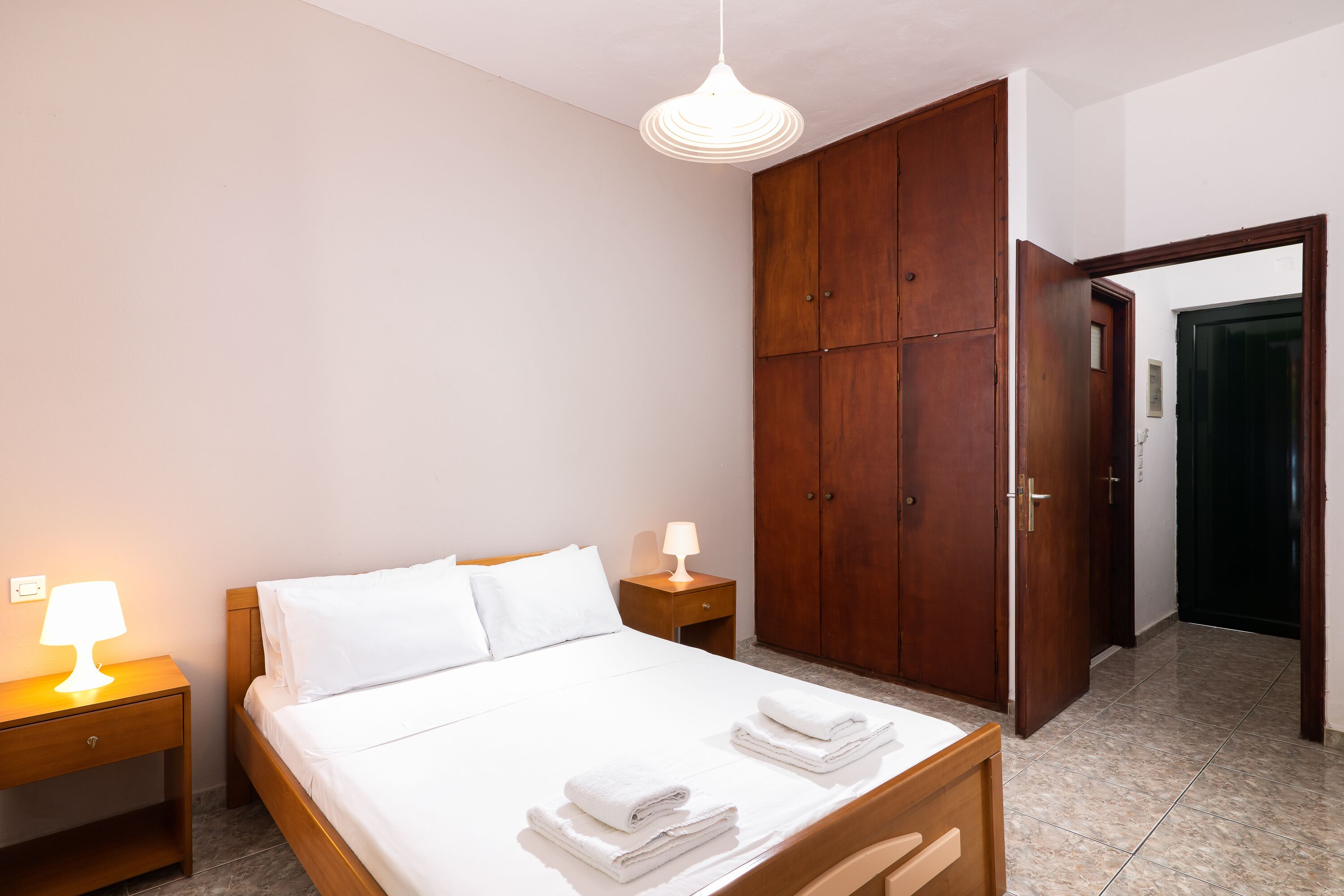 A neatly made bed with white linens and two folded towels, a wooden wardrobe, a bedside table with a lamp, and a door leading to another room.