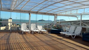 A sunlit deck with white lounge chairs and a blue sofa, large windows offering ocean views, and a glass ceiling structure. - Mar de Canasvieiras Hotel & Eventos (Florianópolis)