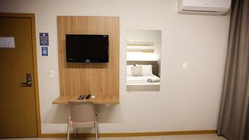 A hotel room with a wooden desk, a television mounted on the wall, a bed with a headboard, and an air conditioning unit.