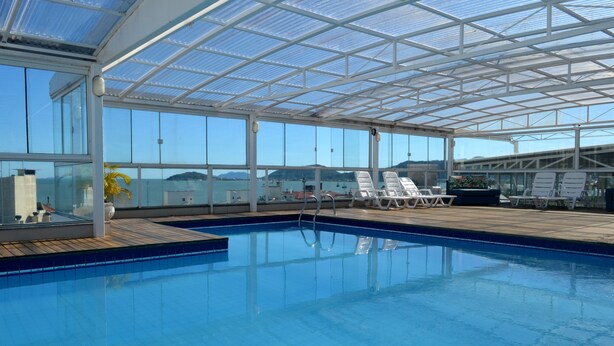 A covered swimming pool with clear glass walls, lounge chairs, and a view of the sea. - Mar de Canasvieiras Hotel & Eventos (Florianópolis)