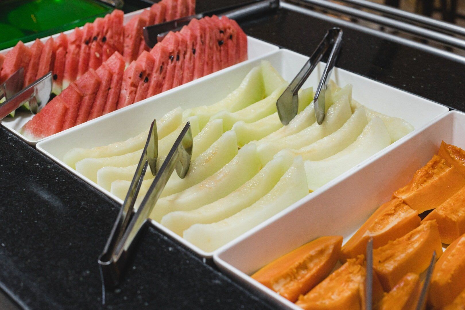 A fruit display with tongs in a white box, containing sliced watermelon, melon, and papaya.