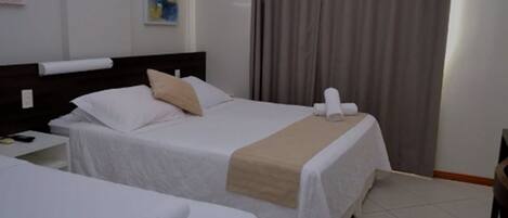 Quarto de hotel com duas camas, cabeceira, mesa de cabeceira, abajur e cortina.