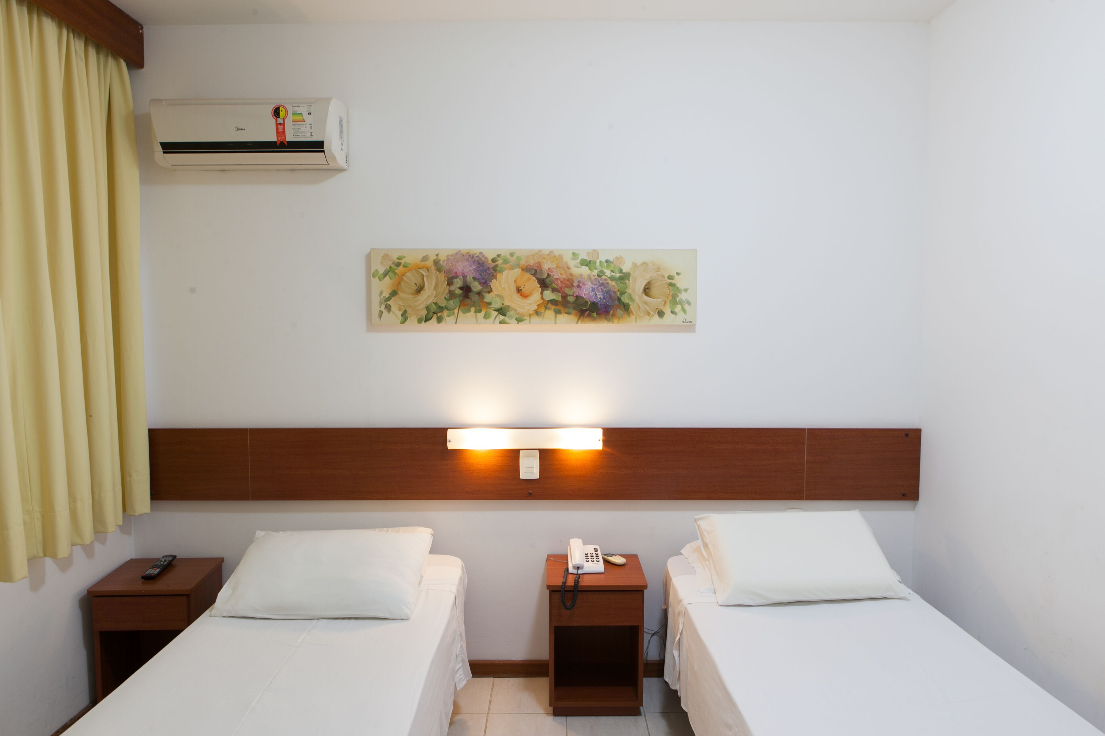 Habitación de hotel con dos camas individuales, una mesita de noche con teléfono, aire acondicionado instalado en la pared y un cuadro sobre las camas.