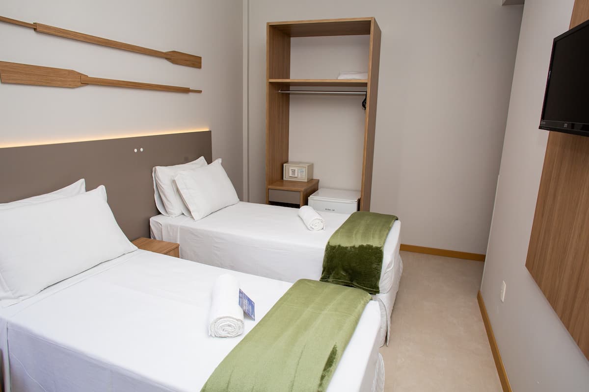 Ein Hotelzimmer mit zwei Betten, einem Holzregal darüber, einem Fernseher an der Wand und einem kleinen Nachttisch mit Lampe.