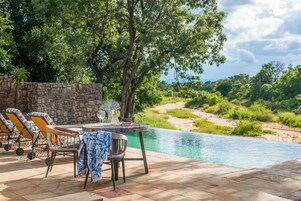 Zona de piscina con sillas y una mesa, rodeada por un muro de piedra y abundante vegetación.