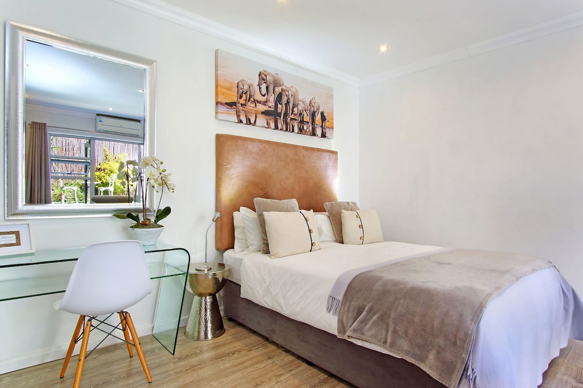 A bedroom with a glass desk, a bed with a headboard, a potted plant, and a painting on the wall.
