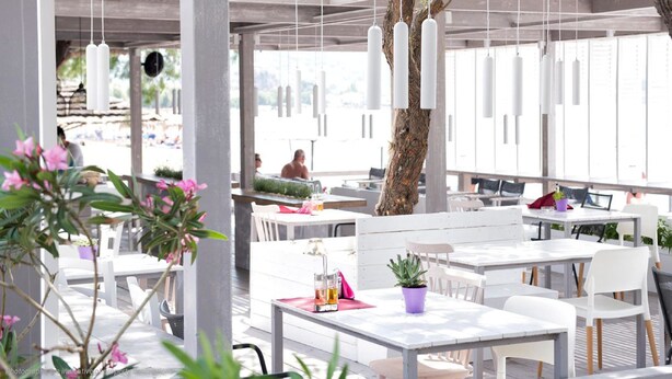 An outdoor seating area with white tables and chairs, hanging pendant lights, and potted plants. - Gaea Gardens Studios (Lesvos)