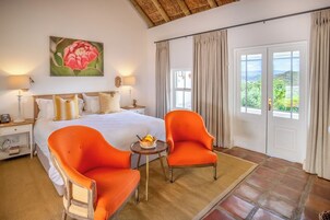 Quarto com cama grande, duas poltronas, mesa pequena com frutas e pintura de flor na parede.