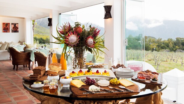 A breakfast buffet with various food items, a vase of flowers, and a view of a vineyard through large windows. - Galenia Estate (Montagu)