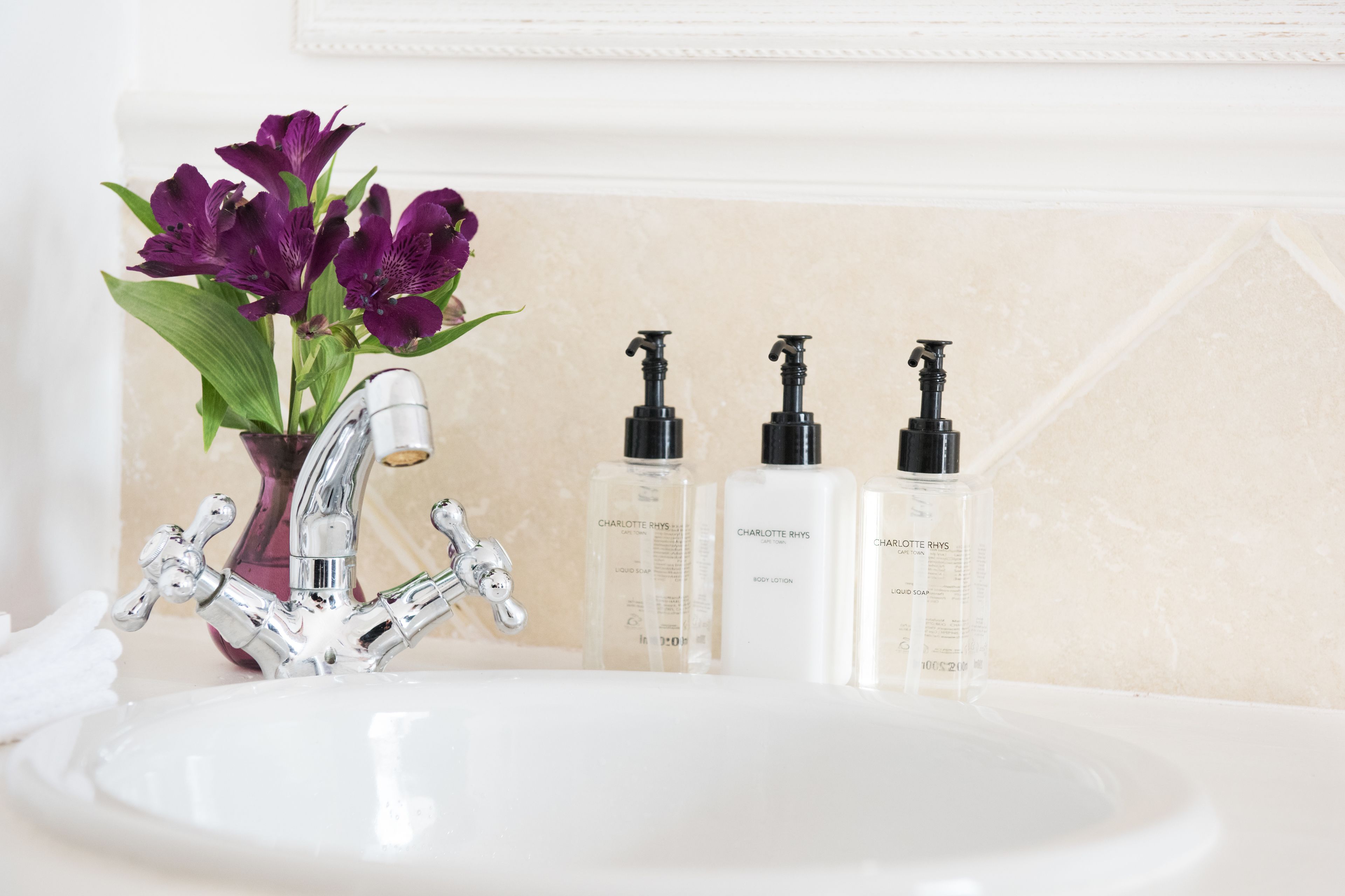 A bathroom sink with a modern faucet, three bottles of Charlotte Rhyss body lotion, and a vase with purple flowers.