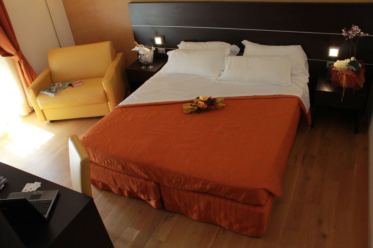 Una habitación de hotel con una cama grande, un sillón amarillo, un escritorio con una computadora portátil y una mesita de noche con una lámpara.
