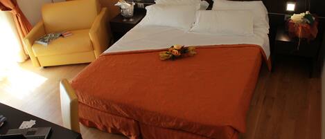 A hotel room with a large bed, a yellow armchair, a desk with a laptop, and a nightstand with a lamp.