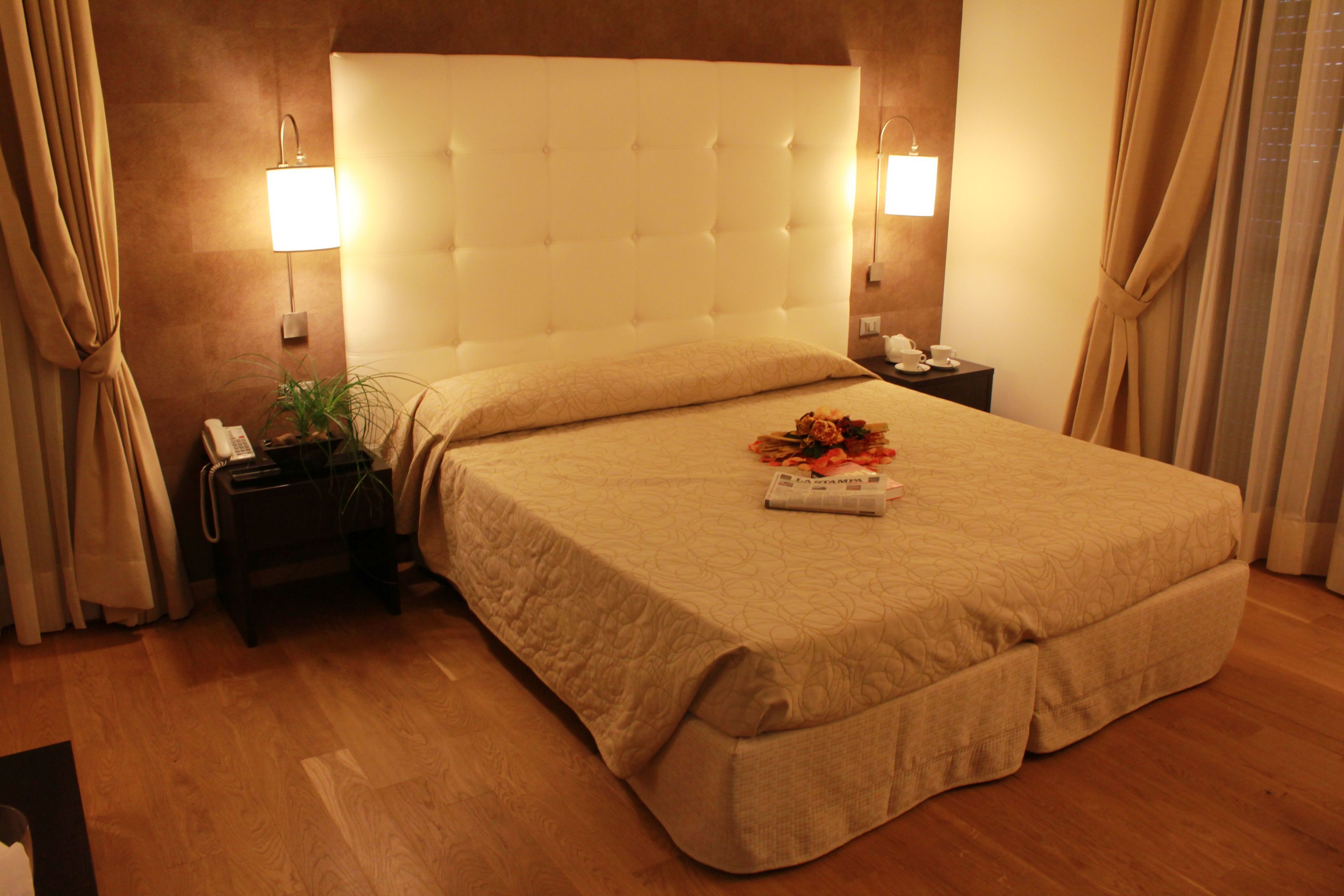 A hotel room with a large bed, a bedside table with a lamp, a nightstand, and a potted plant.