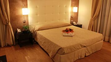 A hotel room with a large bed, a bedside table with a lamp, a nightstand, and a potted plant.