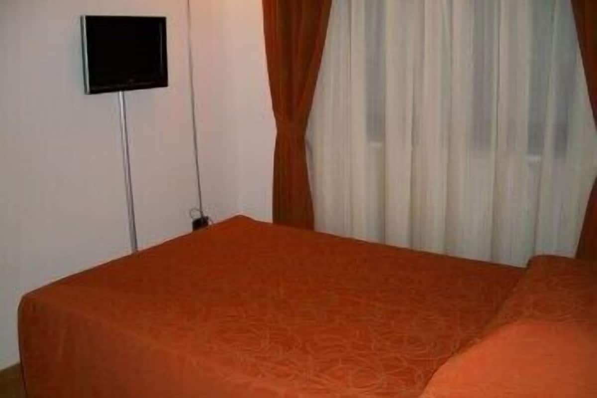 Una habitación con una cama, televisión fijada en la pared y ventana con cortinas.
