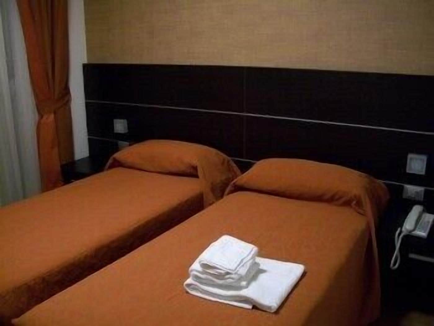 Een hotelkamer met twee enkele bedden, elk met een oranje sprei en passende kussens. Op het bed in de voorgrond ligt een netjes opgevouwen witte handdoek.