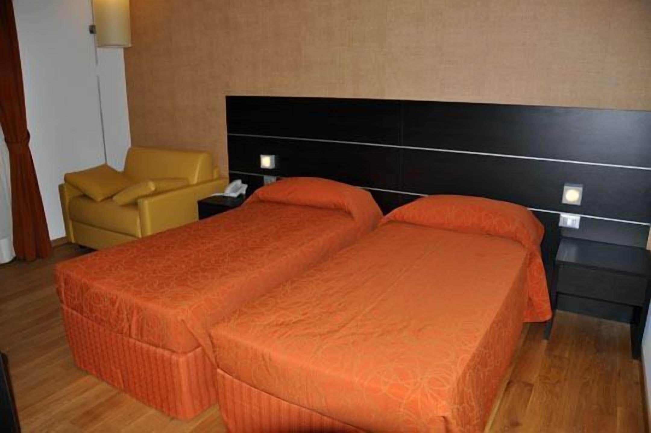 Una habitación de hotel con dos camas, piso de madera, un sillón amarillo y un cabecero negro.