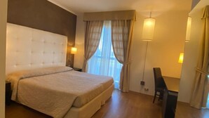 Habitación de hotel con una cama grande, un escritorio y una silla. Se ve una ventana con cortinas y una lámpara sobre el escritorio.
