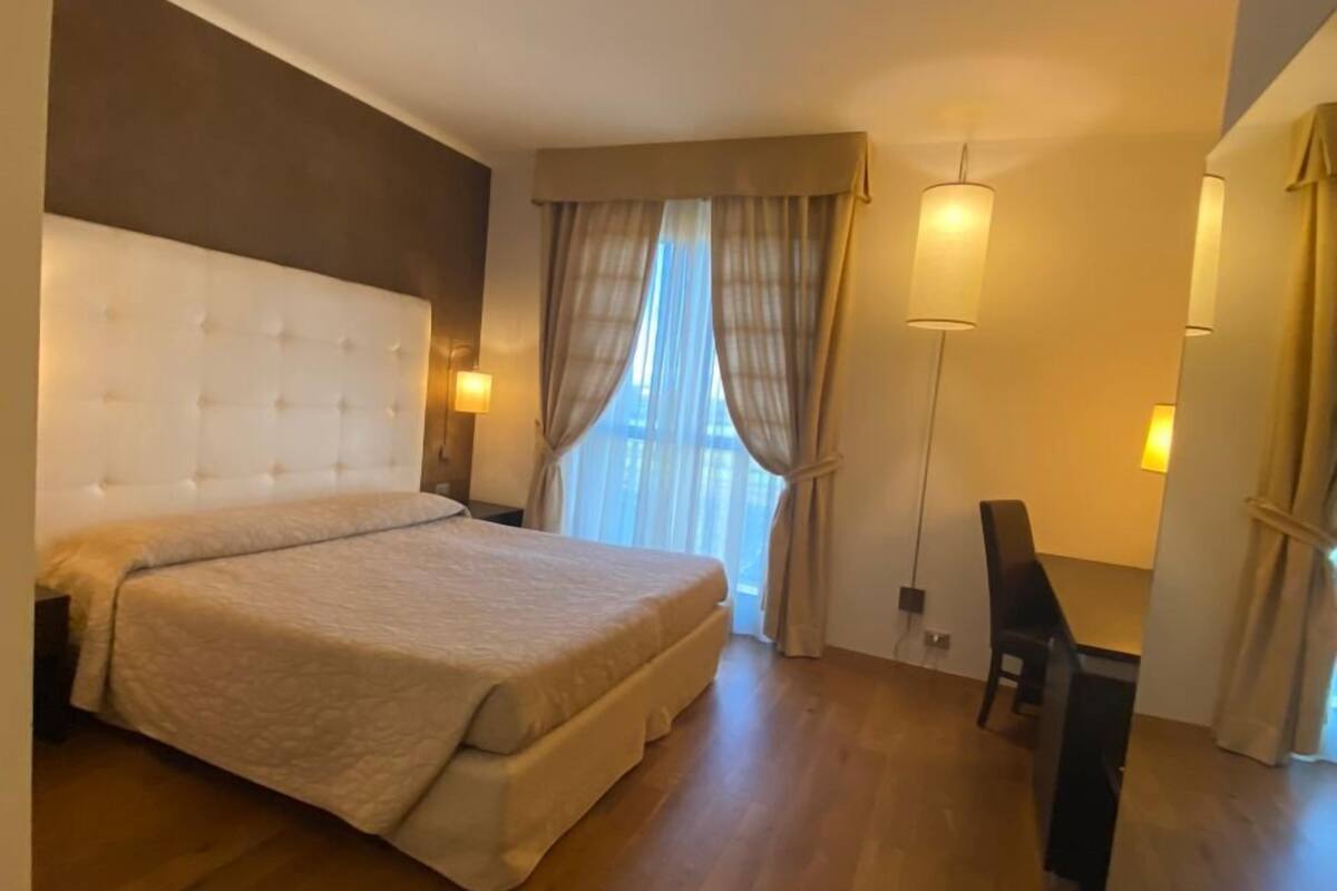 Habitación de hotel con una cama grande, un escritorio y una silla. Se ve una ventana con cortinas y una lámpara sobre el escritorio.