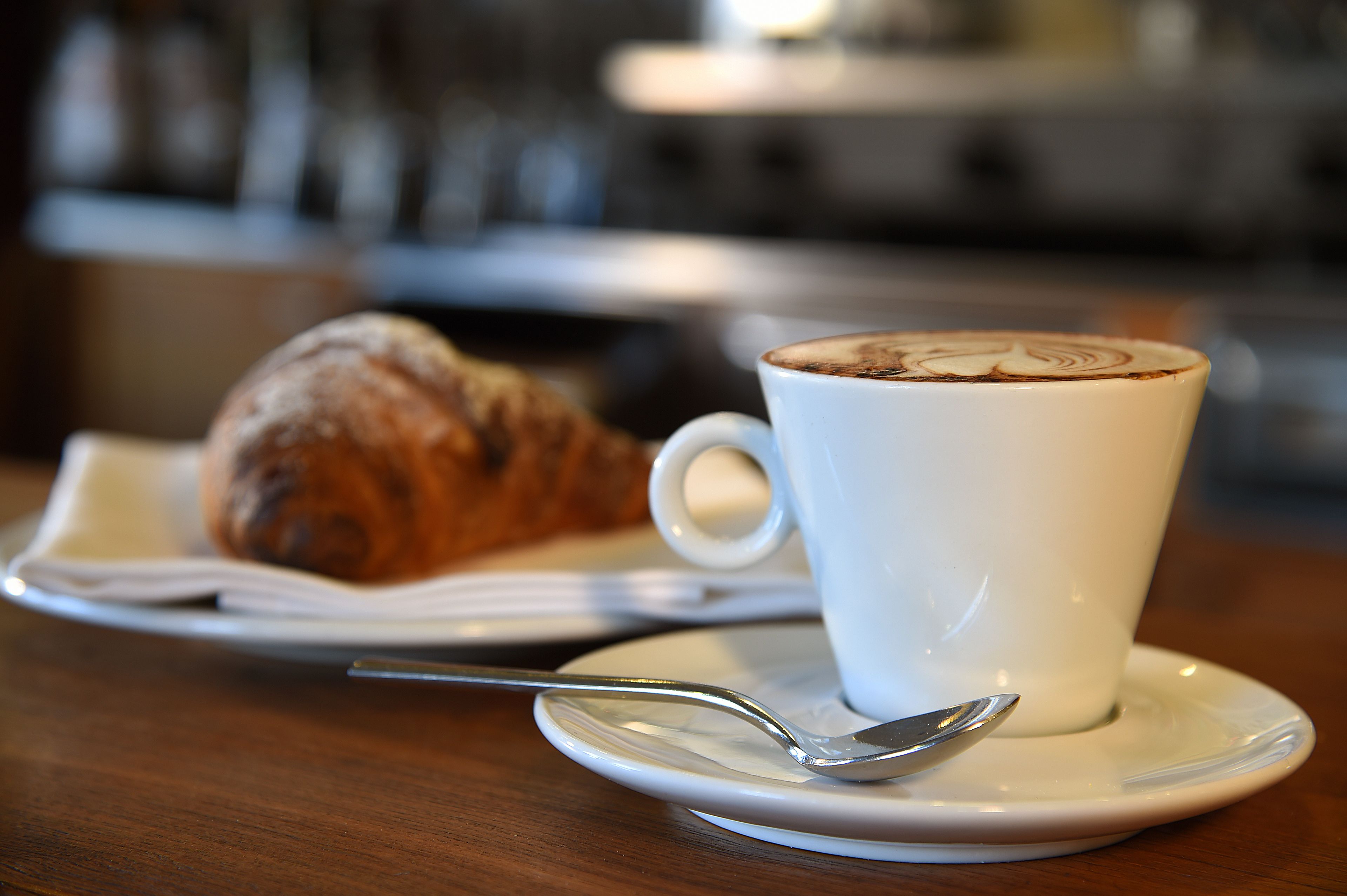 Eine Tasse Kaffee mit Latte-Art auf einem Untertasse, ein Croissant und ein Löffel auf einem Holztisch.