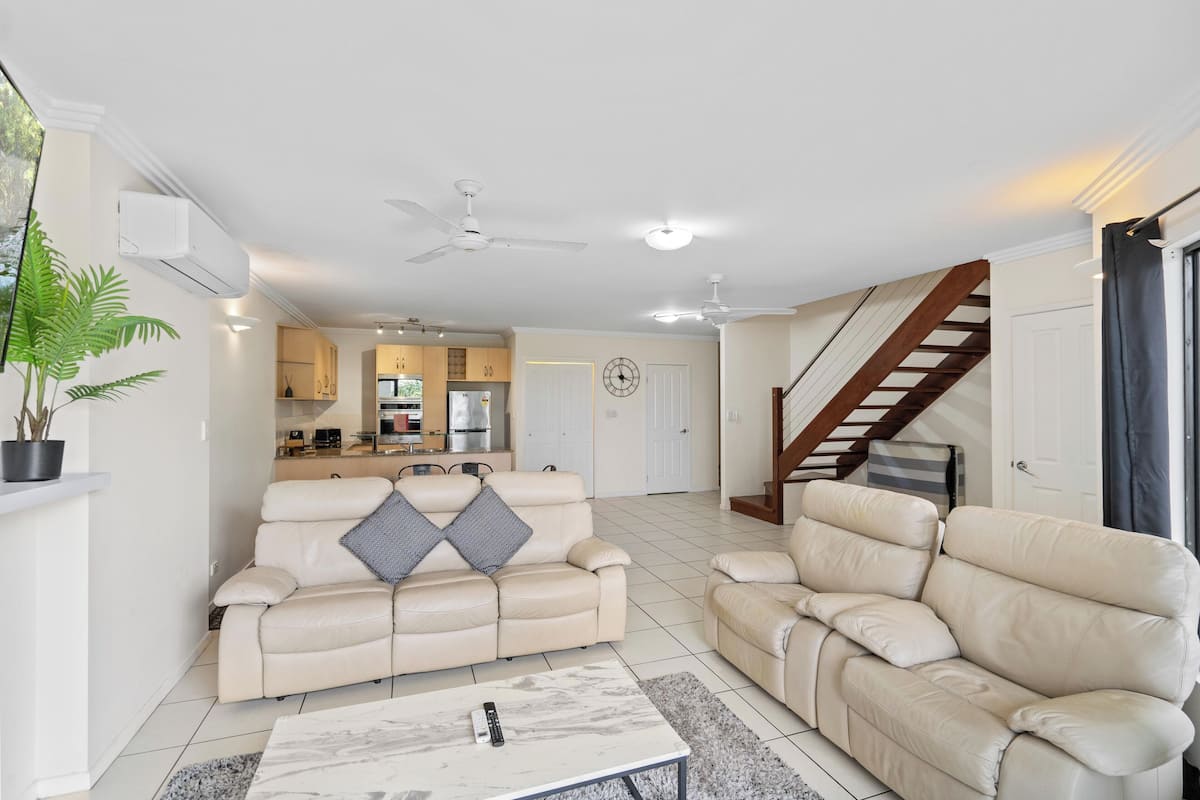 Ein geräumiges Wohnzimmer mit zwei hellbeigen Ledersofas, einem Marmortisch und einer Küche im Hintergrund.