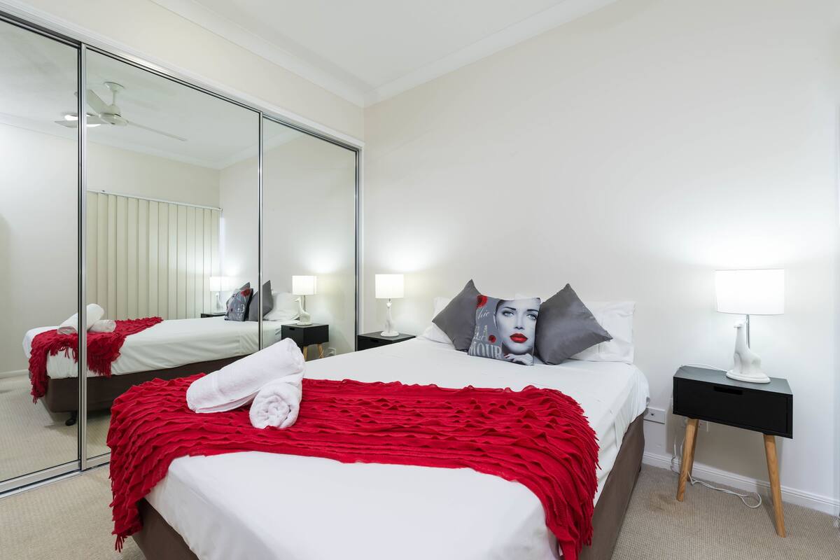 Ein Hotelzimmer mit einem großen Spiegel, einem Bett mit roter Decke, zwei Nachttischen mit Lampen und einem Beistelltisch mit einer Lampe.