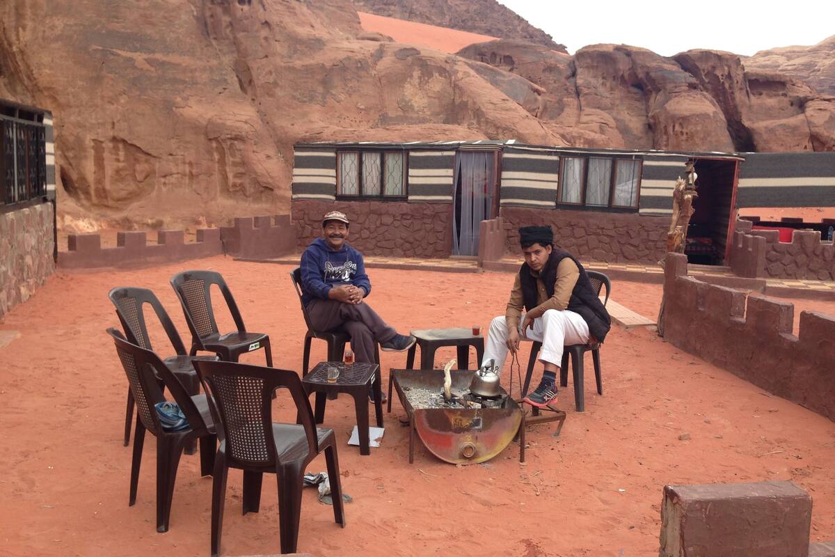Dos hombres sentados a una mesa con sillas, en un paisaje desértico, con un edificio al fondo.