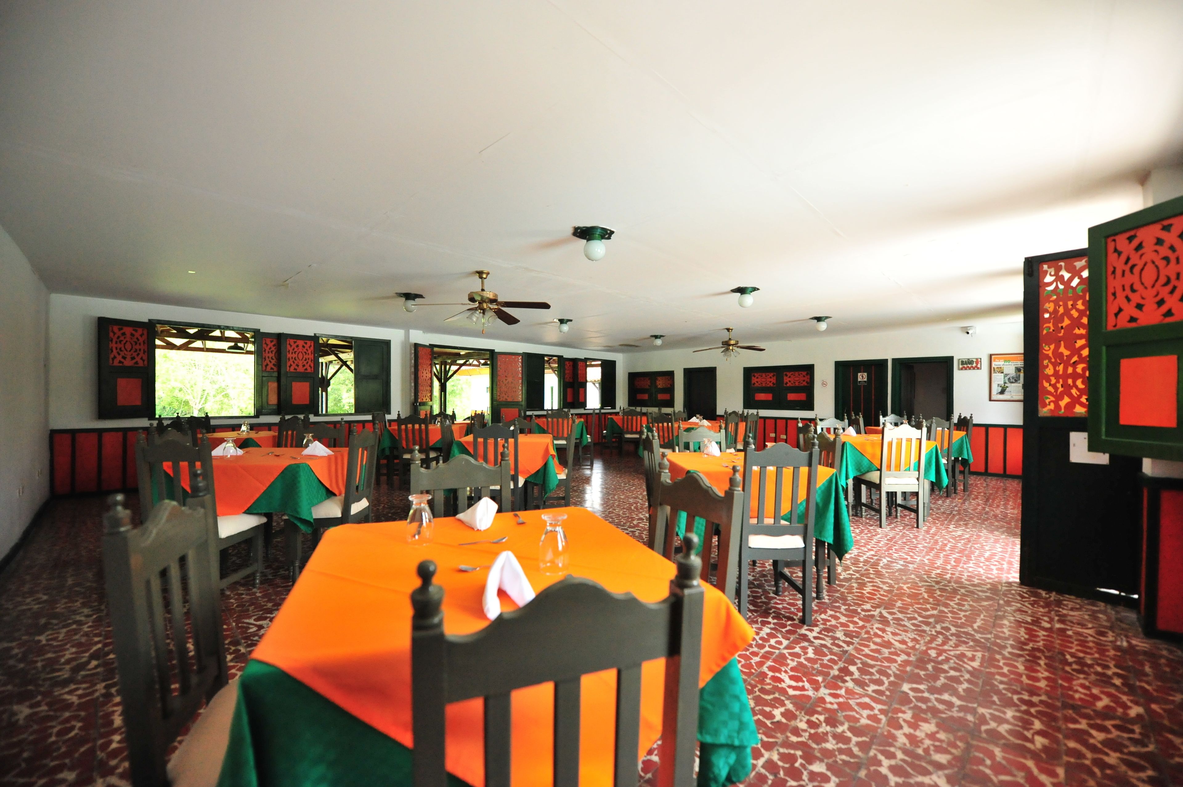 Un espace repas avec des tables recouvertes de nappes orange, des chaises en bois et des ventilateurs de plafond.